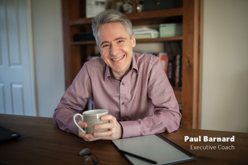 Paul Barnard, executive coach, sitting at a desk with a warm drink.